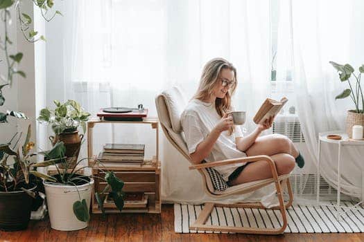 Cheerful young female in eyeglasses with cup of beverage reading textbook in armchair between potted plants in house room