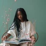 Concentrated young black woman reading illustrated book in green studio near blooming flowers