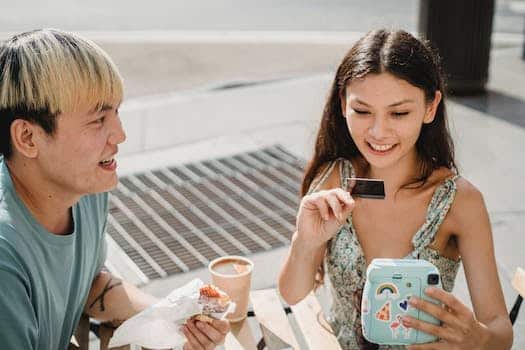 Crop smiling Asian man with cheerful ethnic girlfriend with photo camera at cafeteria table with takeaway coffee