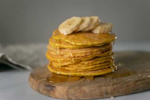 Delicious homemade pancakes covered with honey with sliced bananas placed on wooden chopping board on table in light place