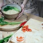 Unrecognizable woman with bowl cooked noodles and leek and unrecognizable person cutting egg on chopping board in kitchen