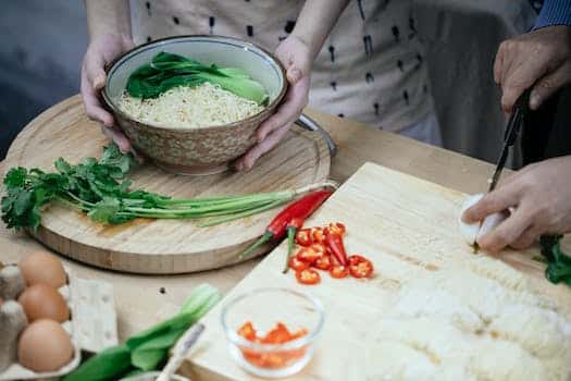 Unrecognizable woman with bowl cooked noodles and leek and unrecognizable person cutting egg on chopping board in kitchen