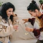 Young happy Hispanic female friends with glasses of champagne having fun sitting on sofa and celebrating holiday