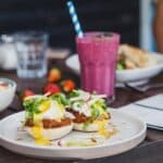 Appetizing sandwiches with poached eggs and sauce served with berry smoothie and placed on table with crop unrecognizable woman reading menu on background