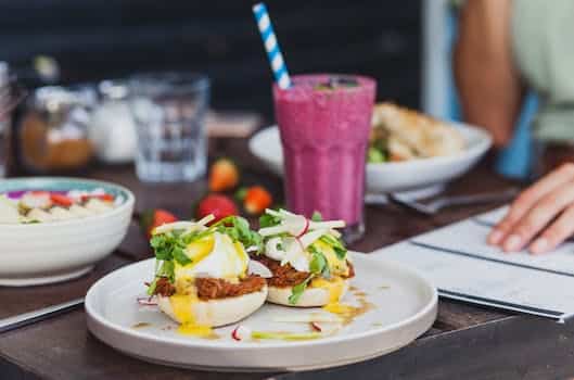 Appetizing sandwiches with poached eggs and sauce served with berry smoothie and placed on table with crop unrecognizable woman reading menu on background