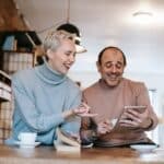 Cheerful adult diverse couple in casual clothes watching funny video on tablet and laughing while drinking coffee at table in apartment