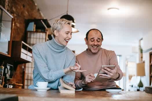 Cheerful adult diverse couple in casual clothes watching funny video on tablet and laughing while drinking coffee at table in apartment