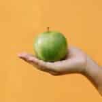 Crop anonymous female demonstrating fruit with thin green skin and crisp flesh against orange background