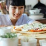 Crop Asian lady smiling and looking at homemade pizza placed on wooden table among various fresh ingredients and utensil in kitchen