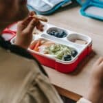 Crop faceless teenage sitting at wooden table with lunch box full of healthy food