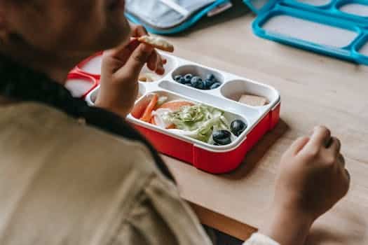 Crop faceless teenage sitting at wooden table with lunch box full of healthy food