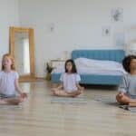 Full body of concentrated multiethnic girls in casual clothing meditating in bright room with eyes closed and arms folded on crossed legs