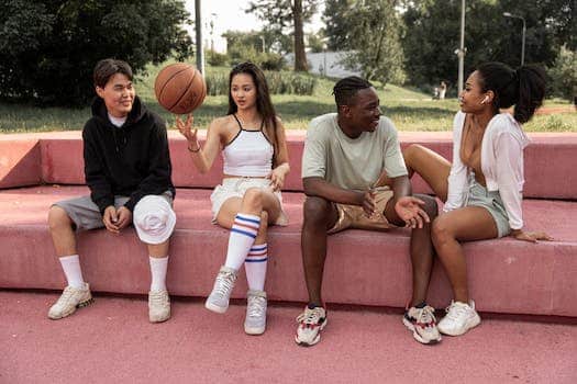 Full length happy young multiracial friends in casual wear sitting on pink stone bench with basketball ball in green park and chatting