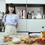 Happy young Asian woman standing near table with ingredients for dough