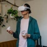 Man in virtual reality headset with controllers playing video game against potted plants in house room