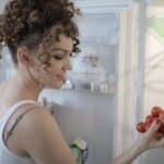 Side view of content female in casual wear standing in kitchen near opened fridge and picking fruit for breakfast