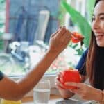 Side view of positive Asian female feeding smiling friend with salad while sitting with cold cocktail in restaurant near window with blurred street