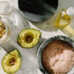 Top view of tasty halved avocado sprinkled with various seeds placed near pink salt with seed mixture in glass pot and other dressing ingredients arranged on white marble table