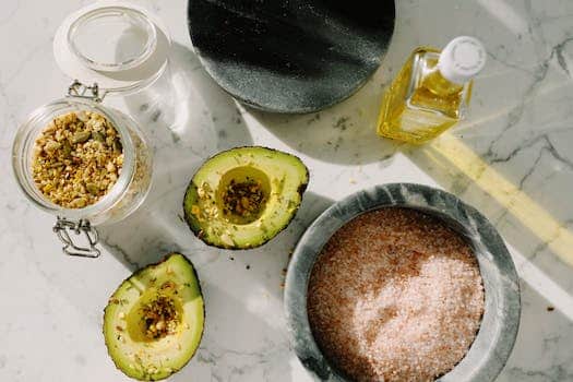 Top view of tasty halved avocado sprinkled with various seeds placed near pink salt with seed mixture in glass pot and other dressing ingredients arranged on white marble table