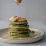 Unrecognizable person pouring honey on stack of appetizing green pancakes topped with bananas and walnuts and served on plate on table