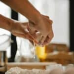 Unrecognizable woman cracking egg into flour on table making dough in kitchen on blurred background