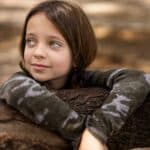 Adorable little girl in casual wear leaning on dry tree trunk in woodland and looking away dreamily