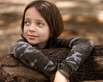 Adorable little girl in casual wear leaning on dry tree trunk in woodland and looking away dreamily