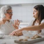 Calm senior woman and teenage girl in casual clothes looking at each other and talking while eating cookies and cooking pastry in contemporary kitchen at home