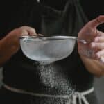 Crop anonymous cook in apron sifting flour while preparing baking dish against gray background