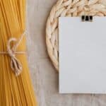 From above of blank paper placed on wicker table mat next to spaghetti tied with bow arranged on table covered with linen tablecloth suitable for recipe or ingredient listing