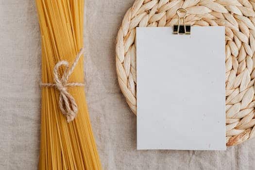 From above of blank paper placed on wicker table mat next to spaghetti tied with bow arranged on table covered with linen tablecloth suitable for recipe or ingredient listing