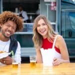 Positive young multiracial friends eating fast food and drinking beer in outdoor cafe