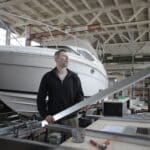 Side view of adult bearded workman in casual clothes standing near workbench and controlling metal detail in garage with yachts