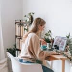 Side view of attentive female remote employee with cup of tea watching photo gallery on netbook screen at desk in house