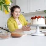 Smiling young female confectioner in casual clothes finishing decoration of tasty cake with chocolate glaze and berries