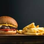 Appetizing burger with meat patty ketchup and cheese placed on wooden table with crispy french fries against black background