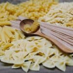 Arrangement of uncooked various pasta including spaghetti fusilli farfalle and penne heaped on table with wooden spoon