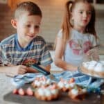 Children in casual clothes near counter with cake and delicious desserts with strawberry and cream in light kitchen