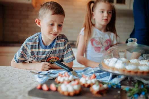 Children in casual clothes near counter with cake and delicious desserts with strawberry and cream in light kitchen