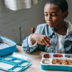 Crop contemplative African American schoolchild looking away at table with lunch container full of yummy food