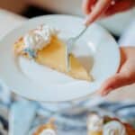 High angle of crop anonymous female cut with fork cake piece with cream on plate near table in bright kitchen