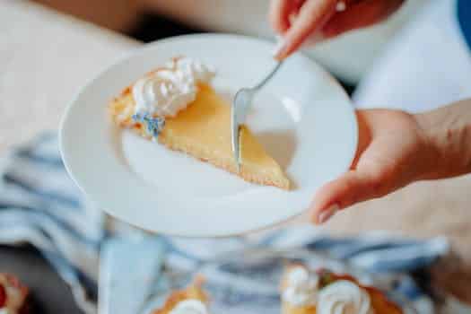 High angle of crop anonymous female cut with fork cake piece with cream on plate near table in bright kitchen