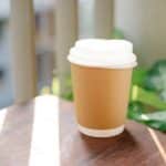 Paper cup of coffee to go placed on table in cafeteria in sunny morning