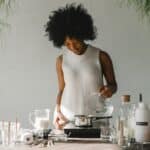 Positive African American female artisan pouring liquid in pot while making wax for candles in pot on cooker