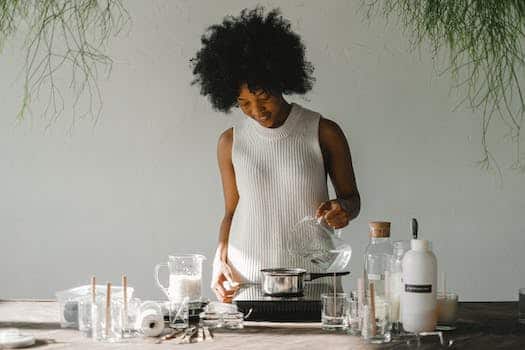 Positive African American female artisan pouring liquid in pot while making wax for candles in pot on cooker