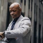 Serious African American businessman in formal suit looking at wristwatch while standing outside modern business center building