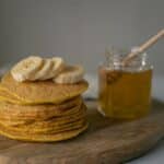 Tasty homemade pancakes with sliced bananas near jar with honey and wooden spoon on cutting board on table