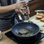 Unrecognizable female cook wearing apron adding seasoning on cutlets while preparing delicious burger at table with stove in kitchen