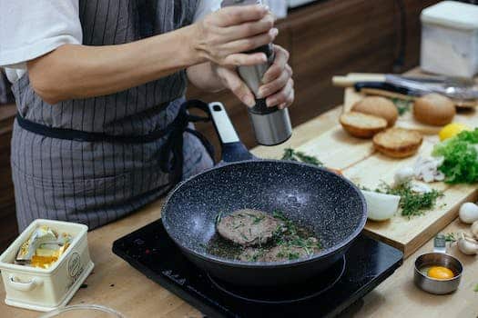 Unrecognizable female cook wearing apron adding seasoning on cutlets while preparing delicious burger at table with stove in kitchen