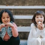 Cheerful multiethnic girls eating tasty appetizing gingerbread made for Halloween celebration while sitting on steps